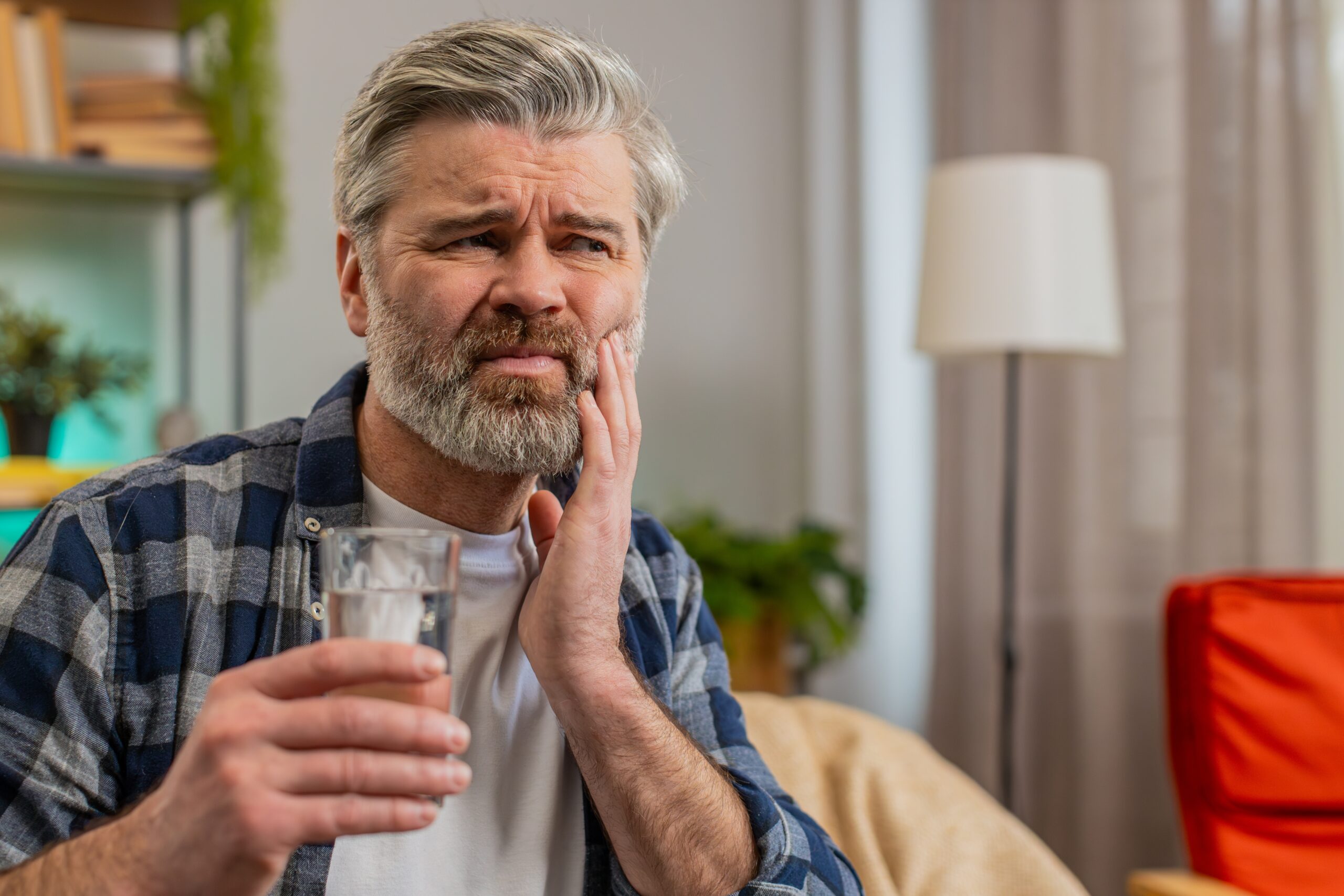 man sitting on a couch with a glass of water holding jaw, broken tooth